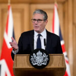 Man in a dark suit and tie speaks at a wooden lectern with a crest, flanked by UK flags in a formal press setting.
