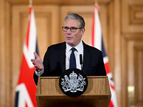 Man in a dark suit and tie speaks at a wooden lectern with a crest, flanked by UK flags in a formal press setting.