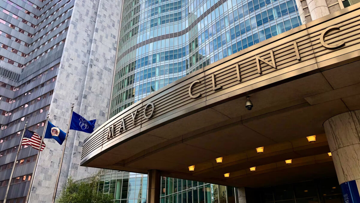 Mayo Clinic building entrance with a curved metal canopy reading MAYO CLINIC, glass facade behind, and American and blue flags on flagpoles.