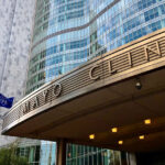 Mayo Clinic building entrance with a curved metal canopy reading MAYO CLINIC, glass facade behind, and American and blue flags on flagpoles.