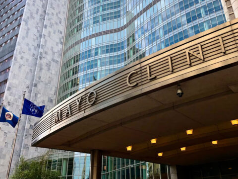 Mayo Clinic building entrance with a curved metal canopy reading MAYO CLINIC, glass facade behind, and American and blue flags on flagpoles.