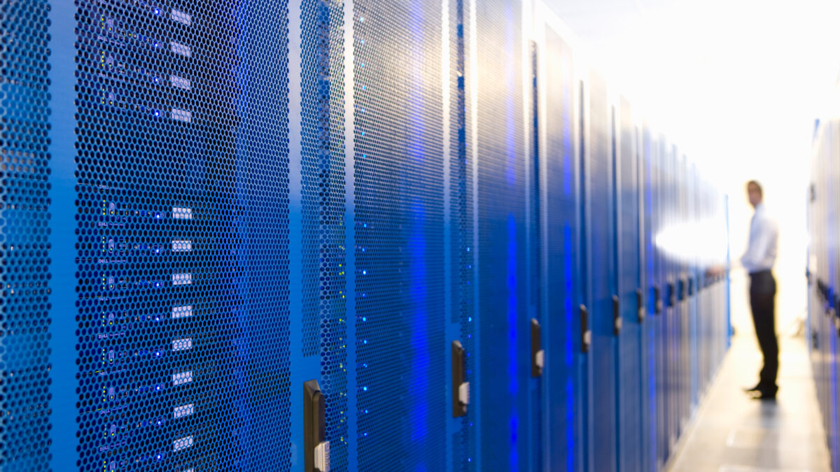 Row of blue server racks with perforated fronts in a data center; a blurred technician stands in the background to the right.