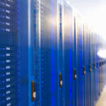 bright-blue-row-of-network-server-cabinets-with-me-2026-01-11-08-42-14-utc - Alpha Row of blue server racks with perforated fronts in a data center; a blurred technician stands in the background to the right.