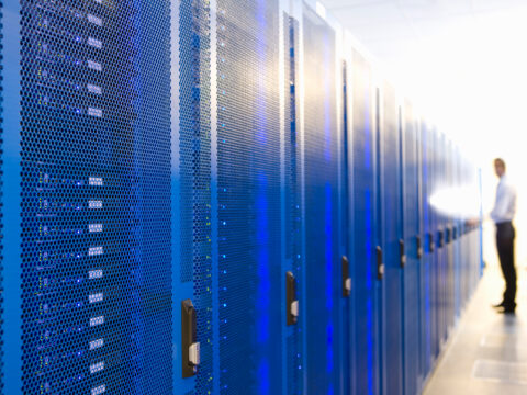 Row of blue server racks with perforated fronts in a data center; a blurred technician stands in the background to the right.
