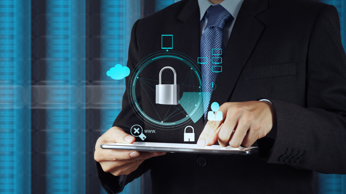 Businessperson in a suit holding a tablet with holographic security icons (padlock, cloud, network) overlay to represent cybersecurity.