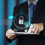 Businessperson in a suit holding a tablet with holographic security icons (padlock, cloud, network) overlay to represent cybersecurity.