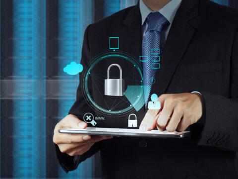 businessman-securing-data-on-tablet-with-digital-g-2026-04-13-23-43-44-utc - Alpha Businessperson in a suit holding a tablet with holographic security icons (padlock, cloud, network) overlay to represent cybersecurity.