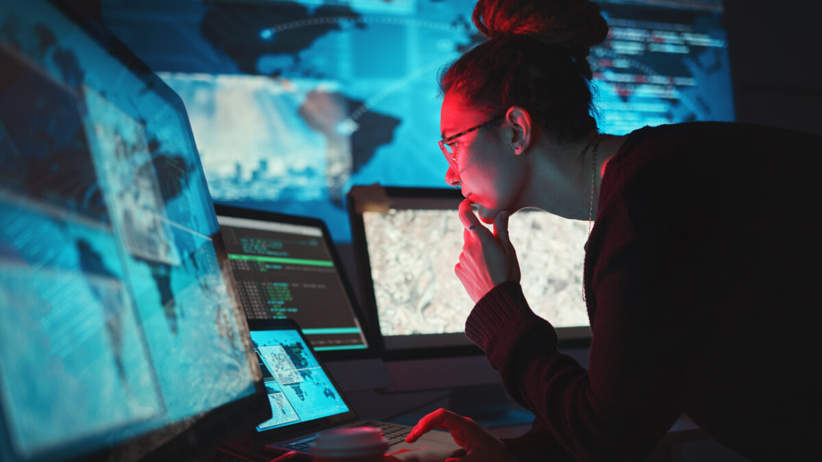 Person with glasses and a bun studies code and maps on multiple screens in a dim blue-lit data room.