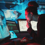 Person with glasses and a bun studies code and maps on multiple screens in a dim blue-lit data room.