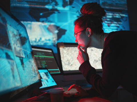 focused-woman-working-with-multiple-computer-scree-2026-03-25-03-30-58-utc - Alpha Person with glasses and a bun studies code and maps on multiple screens in a dim blue-lit data room.