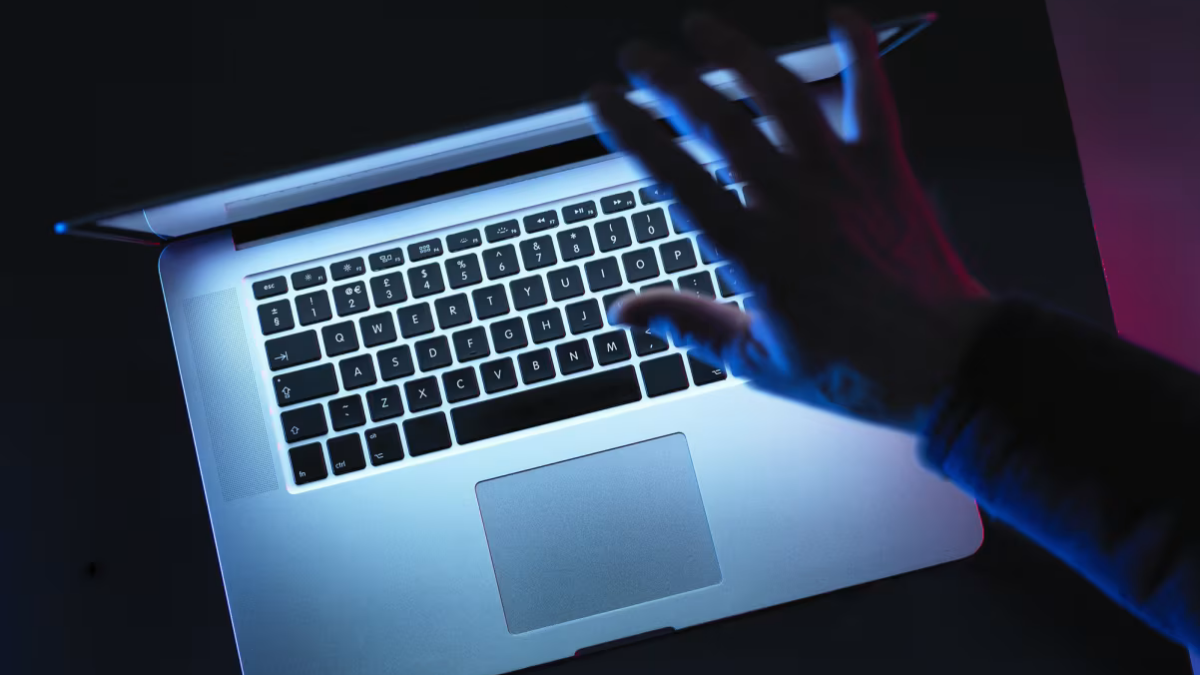 Hand typing on a silver laptop keyboard bathed in blue neon light in a dark setting