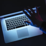 Hand typing on a silver laptop keyboard bathed in blue neon light in a dark setting