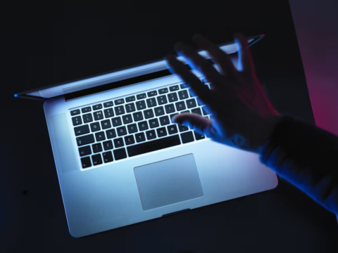 Hand typing on a silver laptop keyboard bathed in blue neon light in a dark setting