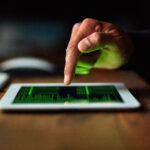 Close-up of a hand tapping a tablet displaying a green data visualization; keyboard blurred in the background.