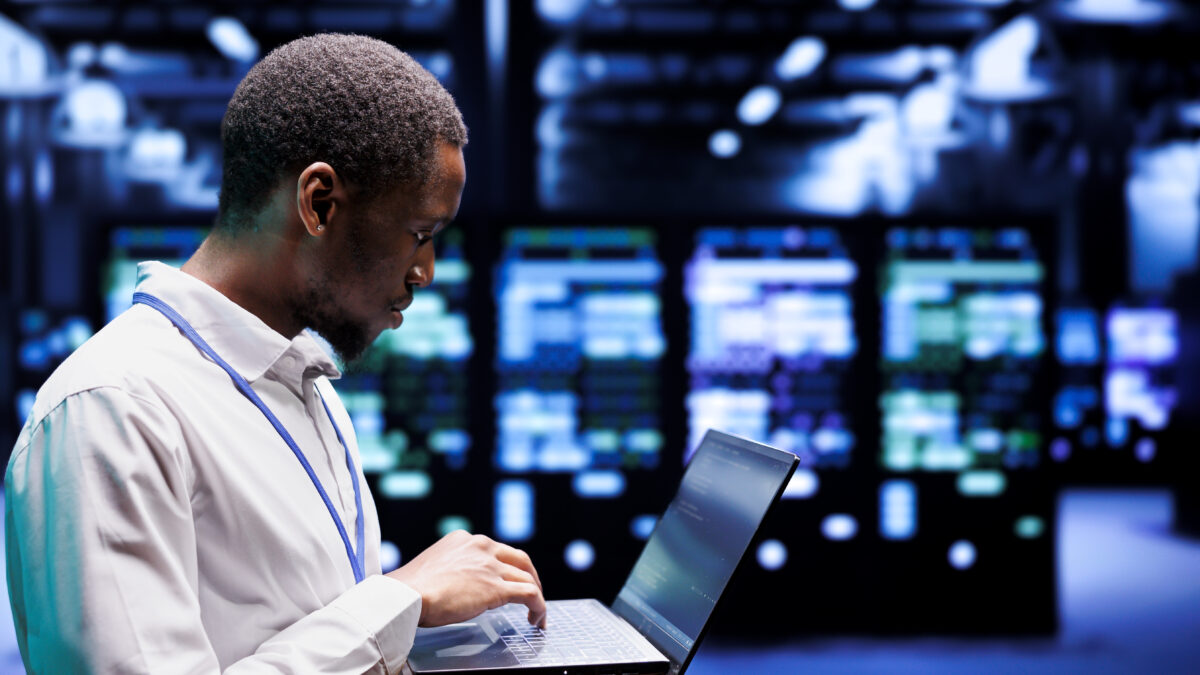 Man in a white lab coat uses a laptop in a server room with blue lighting and rows of equipment behind him.