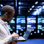 Man in a white lab coat uses a laptop in a server room with blue lighting and rows of equipment behind him.
