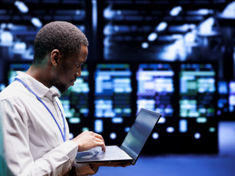 Man in a white lab coat uses a laptop in a server room with blue lighting and rows of equipment behind him.