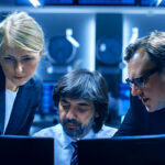 Three professionals in business attire hover over computer monitors in a high-tech control room.