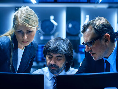 Three professionals in business attire hover over computer monitors in a high-tech control room.