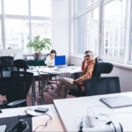 Bright open-plan office with multiple desks, laptops, and a man in an orange hoodie looking toward the camera near large windows.