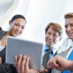 Three business professionals in suits smiling as they review content on a tablet together in a bright, modern environment.