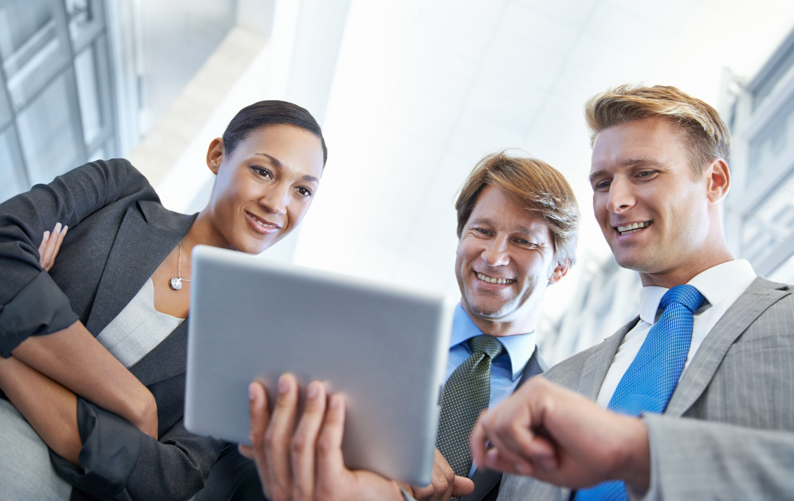 Three business professionals in suits smiling as they review content on a tablet together in a bright, modern environment.