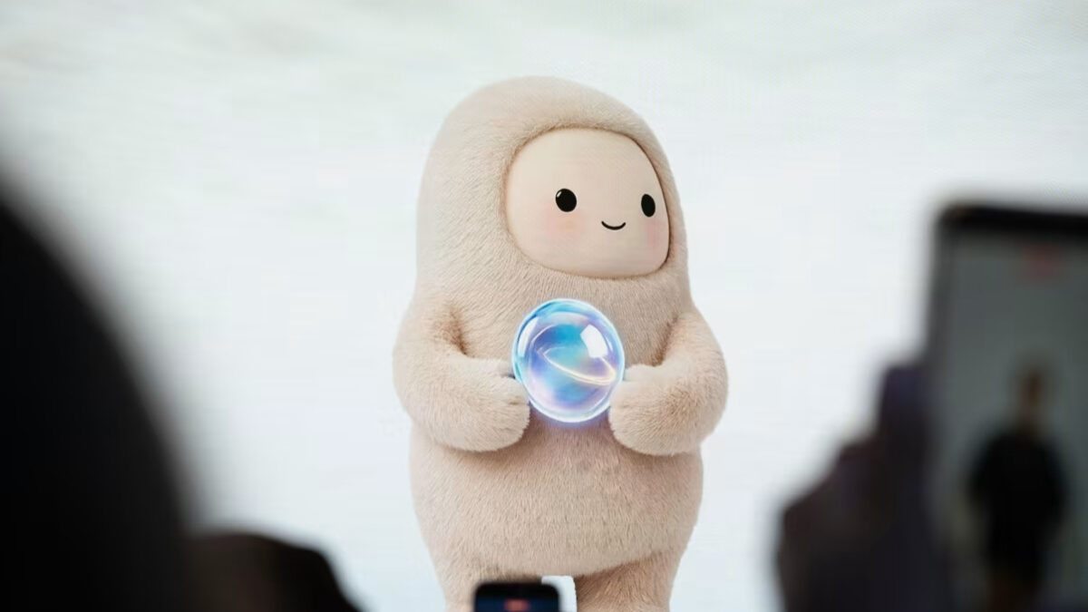 Soft beige mascot in a hooded suit on stage, holding a glowing bubble, audience blurred in the foreground.