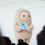 gettyimages-2296214136 - Alpha Soft beige mascot in a hooded suit on stage, holding a glowing bubble, audience blurred in the foreground.