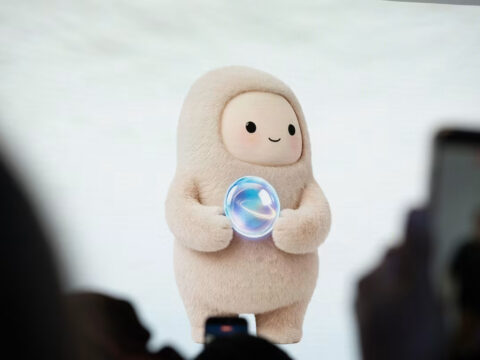 Soft beige mascot in a hooded suit on stage, holding a glowing bubble, audience blurred in the foreground.
