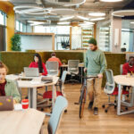 Open-plan coworking space with people at desks; a man in a green beanie stands beside a bicycle among round tables and modern chairs: bright, collaborative office.