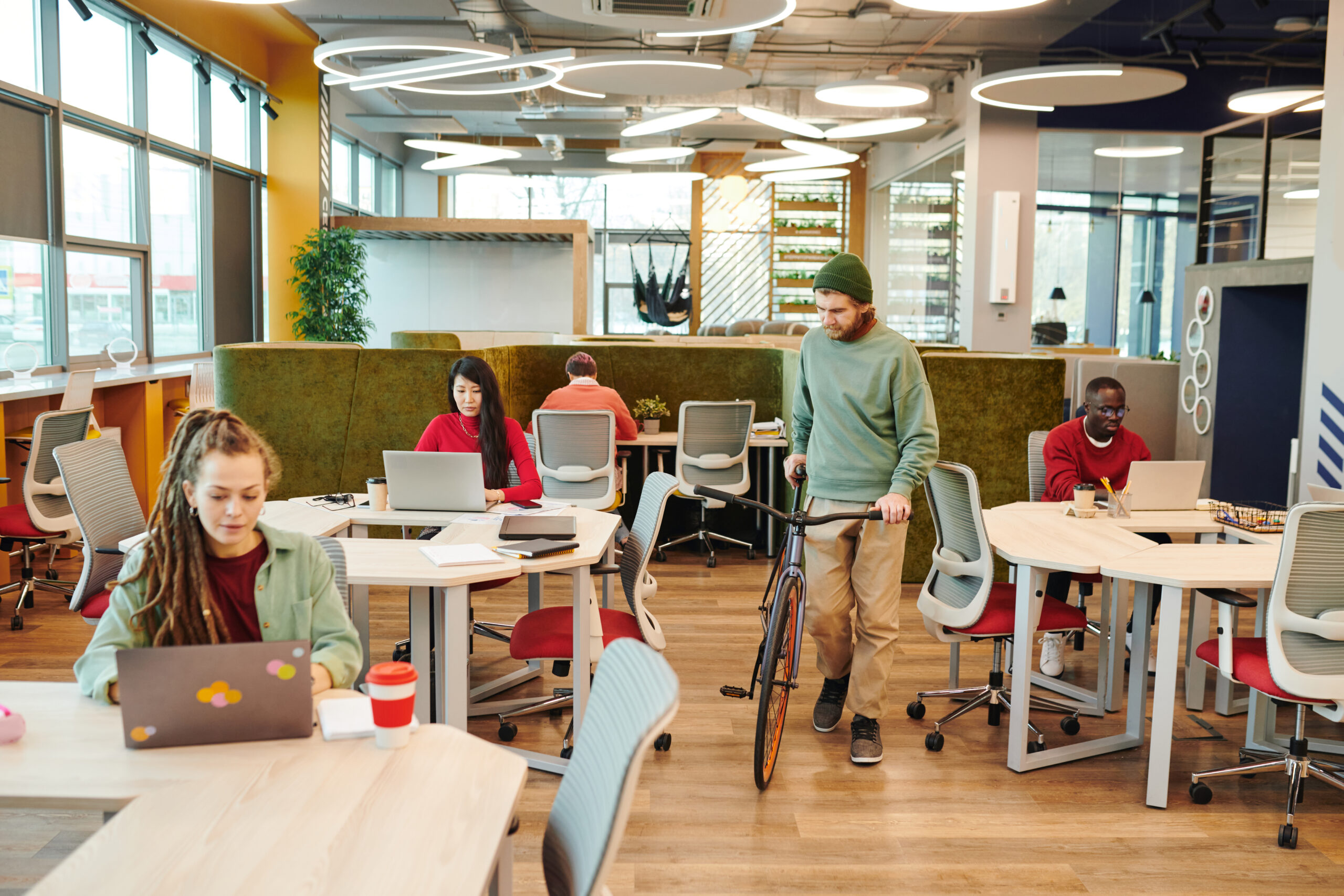 Bearded office manager holding bicycle while moving between desks of colleagues - Alpha Open-plan coworking space with people at desks; a man in a green beanie stands beside a bicycle among round tables and modern chairs: bright, collaborative office.