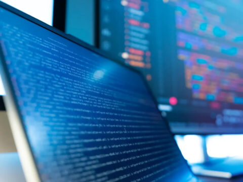 GettyImages-2263067296_2 - Alpha Close-up of a laptop screen filled with blue lines of code, with a second monitor showing colorful data in the blurred background.