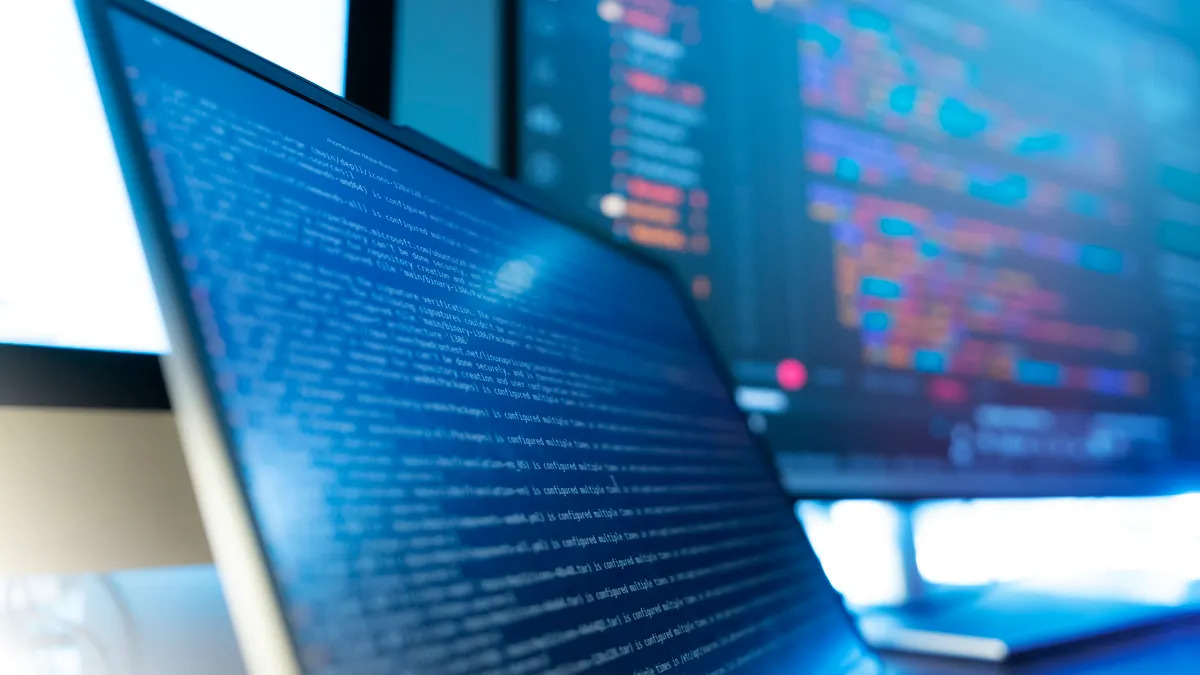Close-up of a laptop screen filled with blue lines of code, with a second monitor showing colorful data in the blurred background.