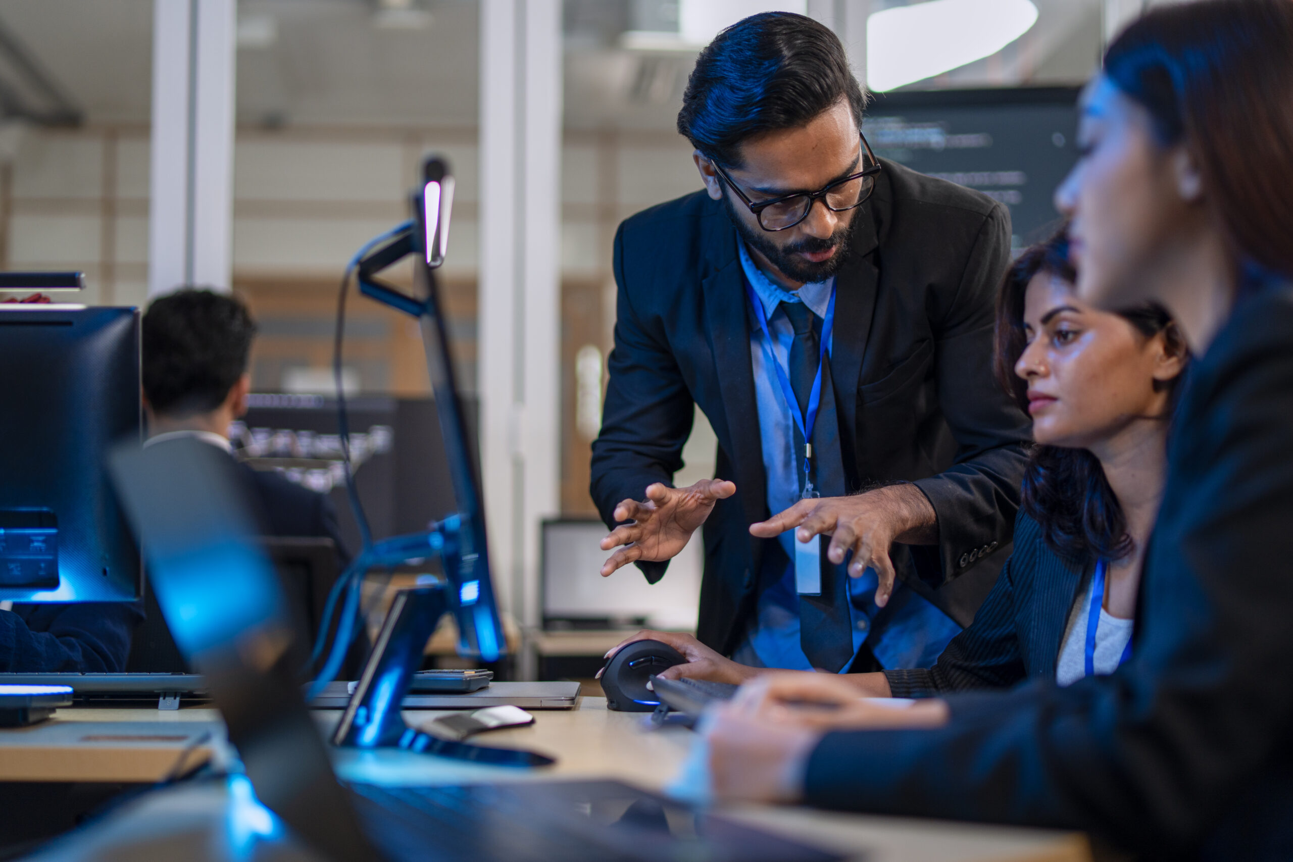 Bearded man in a suit and glasses leans over a desk, gesturing as colleagues work at computers in a tech office study area